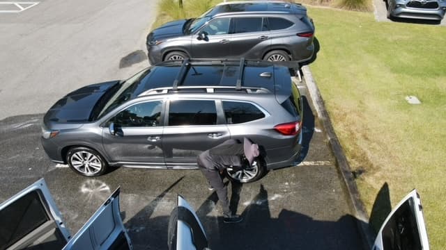 Person in a hat cleaning the wheel of a gray SUV in a parking lot.